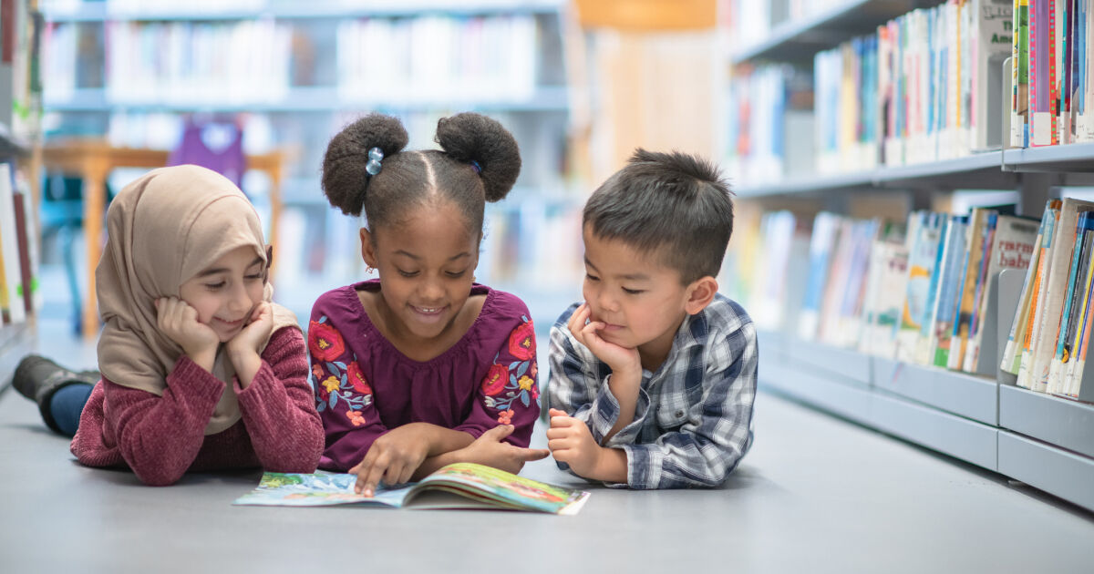 Three young children - a girl in a hijab, another girl with two pigtails in her hair, and a boy wearing a plaid shirt - sit on the floor in a library. They all read the same book in front of them.